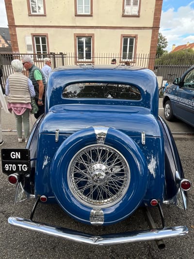 The image depicts a dark blue vintage coupe, viewed from the rear, parked on asphalt pavement in Molsheim, France. The vehicle features a rear-mounted spare wheel with a blue cover and chrome wire spokes, a chrome rear bumper, and circular red taillights. A black license plate displaying "GN" and "970 TG" is affixed to the left rear. An exhaust pipe extends from beneath the right side of the vehicle.

In the background, behind a black metal fence, an elderly woman in a beige quilted vest and light green pants stands facing an elderly man in a green polo shirt and dark vest. Other indistinct individuals are present further back. To the right of the vintage car, a dark blue vehicle with "GENDARMERIE" text in white is partially visible. A blue bicycle is also discernible behind the fence. The primary background structure is a two-story building with a light beige facade, reddish-brown trim, and multiple white-framed windows, some with closed white shutters. The sky above is partly cloudy.