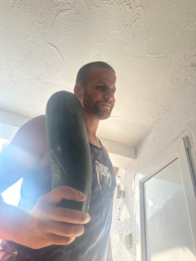 A man, approximately 40-50 years old, with short brown hair and a beard, stands indoors, holding a large zucchini in his right hand. He is wearing a dark-colored tank top with a graphic print. The background features a textured white ceiling and wall, and a white door frame. Sunlight streams in from the left, creating lens flare and illuminating the scene. The image appears to be taken from a low angle, looking up at the man. The environment suggests a domestic setting, possibly a home or a covered outdoor area, in Heinebach, Germany.