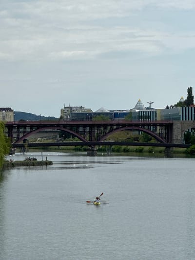 The image shows the Drava River in Maribor, Slovenia, with the Old Bridge (Stari most) spanning the river. The bridge is a metal structure with arched supports. Modern buildings with pyramidal roofs are visible behind the bridge. A single scull rowing boat with a person is on the river's surface. The sky is overcast.