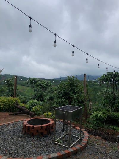 The image depicts an outdoor barbecue area at a rural location, likely in Thôn 4, Vietnam. The scene is characterized by a cloudy, overcast sky that casts a diffused light over the landscape. In the foreground, a circular brick fire pit, filled with grey gravel, is situated next to a metal barbecue grill. A weathered wooden log lies on the gravel nearby. Surrounding the barbecue area is lush green vegetation, including trees and bushes, with a backdrop of rolling hills covered in agricultural terraces, suggesting a coffee or tea plantation. A string of decorative light bulbs hangs across the scene, suspended from a wire, adding to the ambiance of the outdoor space. The overall impression is one of a serene, natural setting, prepared for outdoor gatherings.