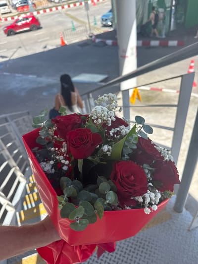 In Mai Khao, Thailand, a bouquet of red roses and baby's breath is held by a person whose arm and shoulder are visible in the lower left. The bouquet is wrapped in red paper. In the background, a woman with long dark hair, wearing a white dress, walks up a set of stairs. Beyond her, a red car is parked on a street with orange traffic cones and white and red striped barriers. The scene appears to be outdoors, possibly near an airport or transportation hub, given the presence of vehicles and barriers.