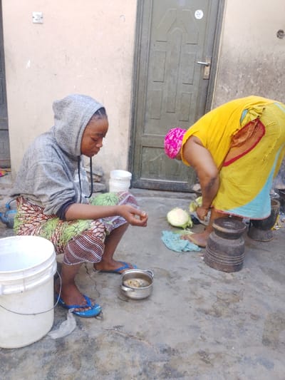 Two women are outdoors preparing food. One woman is seated, wearing a grey hoodie and a colorful skirt, and is focused on something in her hands. A small pot sits nearby. The other woman is bent over, wearing a yellow top and a pink headscarf. She appears to be chopping vegetables with a knife. Wooden tools, including a mortar and pestle, sit next to her. They are in a courtyard-like area near a door.