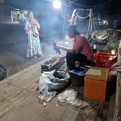 The scene takes place in a street in Kecamatan Asembagus, Indonesia, at night. A man in a pink shirt and black hat sits on a low stool. He is using a metal spatula to stir food cooking on a small, portable grill. A woman in a blue and white dress and a headscarf stands behind him. The street is paved with gray concrete. There is a large gray sack in the foreground. A small, orange cooler is in the lower right corner of the image. There are other objects in the scene, including a plastic container, a metal grill, and several pieces of wood. There is also some smoke coming from the grill. The woman appears to be looking at the man while holding a phone in her right hand. The man is focused on the grill, with smoke and steam in the background obscuring a clear view of his face. The location is determined based on the provided coordinates.