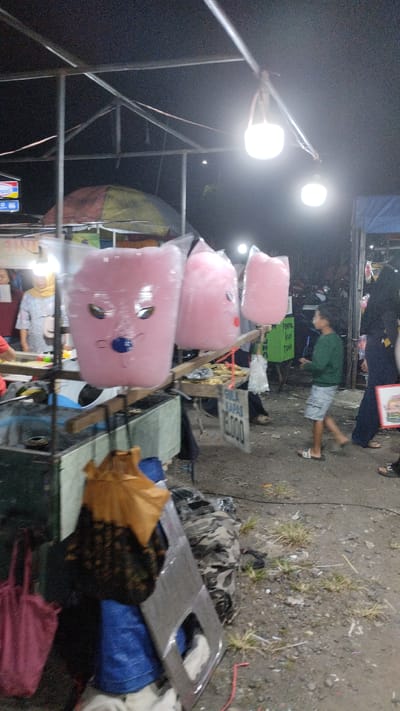 The image depicts a nighttime outdoor market scene, likely in Sambungrejo, Indonesia. The foreground is dominated by a stall selling large, pink, cartoon-faced cotton candy in plastic packaging. To the left of the cotton candy display, a person wearing a yellow hijab is visible, seemingly tending to the stall. Behind the stall, there are umbrellas and market structures. In the mid-ground, a young boy in a green shirt and shorts is walking past. To the right, a person in dark clothing is walking away from the camera. The ground is a mixture of dirt and gravel, with some sparse vegetation. In the background, more market stalls and people are faintly visible under artificial lighting. The overall atmosphere suggests a lively local gathering or event.