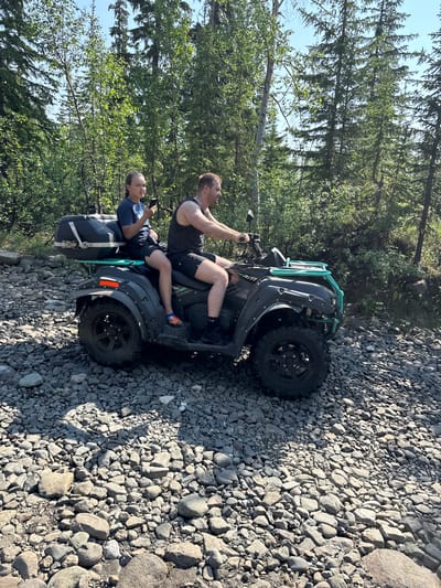 The image shows two people on a quad bike in a rocky, outdoor setting. The person in the front is driving, and the person behind them is looking at a phone. The quad bike is dark gray with a teal roll cage. There are trees in the background, suggesting a natural environment. The ground is covered in gray and brown rocks. The lighting suggests it is daytime, possibly late afternoon, with sunlight casting shadows. The overall mood appears relaxed and casual. There is no visible text in the image.