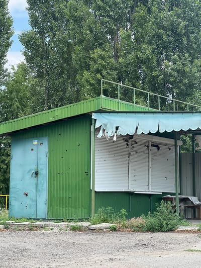 An outdoor image depicts a damaged, single-story, green-sided building or kiosk in Staryi Saltiv, Ukraine. The structure features a corrugated green metal roof with a rusted metal railing along its top edge. The left side of the building has a light blue metal door, secured by a black padlock, showing multiple impact holes consistent with bullet or shrapnel damage. A yellow and blue sticker, resembling the Ukrainian flag or coat of arms, is affixed to the upper left of this door. The green corrugated siding to the right of the door also exhibits numerous impact holes.
The right side of the building has a light blue scalloped awning, heavily torn and frayed. Below the awning are two white horizontal roller shutters, both visibly riddled with multiple impact holes and showing internal damage. To the immediate right of the structure, a brown wooden table and bench are partially visible, covered by a white sheet or tarp. The ground in the foreground is composed of light grey gravel and dirt, with small patches of green weeds and grasses growing near the base of the building. In the background, numerous tall, slender trees with dense green foliage are visible under a partly cloudy sky. No people are present in the scene.