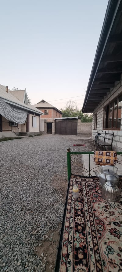 The image depicts an outdoor scene in the city of Jomi, Tajikistan, with a focus on a yard area. In the foreground, a patterned rug is spread over a surface, with two silver teapots and a baby bottle visible on it. A green metal railing borders the rug. The yard is covered in gravel. In the middle ground, there are several buildings. To the left, a light-colored building with a white window is partially visible, with a corrugated metal covering extending from its roof. A dark metal gate with an ornate pattern is situated in the center, separating the gravel yard from another area. Behind the gate, a two-story brick building with dark-trimmed windows is visible. To the right, a white brick building with dark-trimmed windows runs along the edge of the frame, with a black bicycle and a wooden bench placed outside it. The sky is clear and light blue, suggesting it is either early morning or late afternoon.