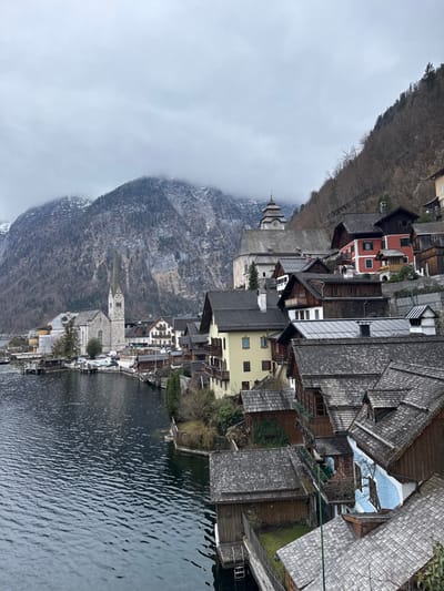 The image displays a detailed view of the village of Hallstatt, Austria, positioned along the shore of Lake Hallstatt against a backdrop of steep, forested mountains. The sky is uniformly overcast with gray clouds, obscuring the upper portions of the rugged, snow-dusted mountain peaks.

In the lower-left and central foreground, the dark, rippled surface of Lake Hallstatt extends. Along the lake's edge, numerous traditional multi-story structures with dark, often shingled roofs are visible. Some buildings, particularly in the foreground, feature small boathouses or extensions built directly over the water.

To the left, a prominent stone church with a tall, light-colored spire (the Evangelical Church) stands close to the lake. Higher on the hillside, to the right of the spire, another distinct stone church with a tiered, dark-roofed tower capped by an onion dome-like structure (the Catholic Church) is visible.

The residential and commercial buildings are densely packed, ascending the steep hillside. Architectural styles include traditional Alpine chalets and townhouses. Facades exhibit a range of colors, predominantly earth tones like creams, yellows, and browns, with notable instances of red and light blue siding. Multiple buildings feature wooden balconies. Sparse, bare deciduous trees and evergreen conifers cover the steep slopes behind and between the structures. No individuals are present in the scene, and no specific actions or interactions are depicted.