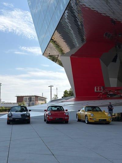 In Stuttgart, Germany, three Porsche automobiles are parked on a paved surface in front of a modern building with a red and silver facade. From left to right, the cars are a black convertible, a red convertible, and a yellow coupe. A man wearing a striped shirt and shorts stands to the right of the yellow car, looking at his phone. In the background, a construction lift is positioned near the building. The sky above is partly cloudy.