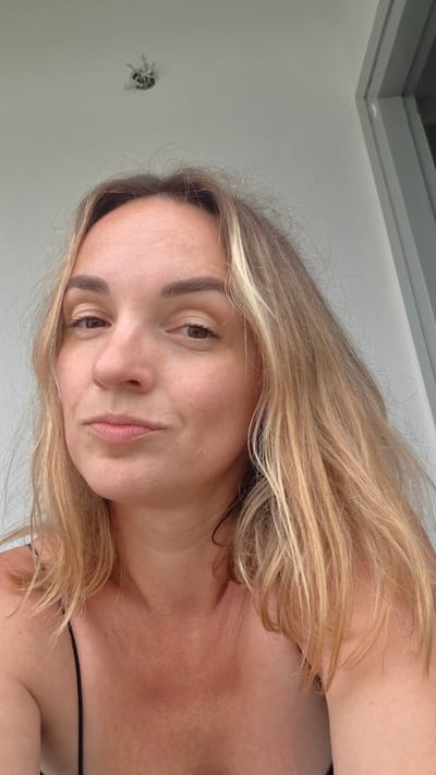 A medium shot captures a woman with blonde hair and brown eyes looking at the camera. She is wearing a black spaghetti strap top. Her skin is lightly tanned and has a few freckles. The background is a white wall with a light fixture at the top. The lighting appears to be natural. The image was taken outdoors, possibly on a balcony, given the presence of a railing visible on the right side of the frame.