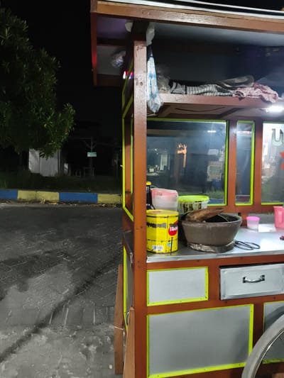 An outdoor, nighttime scene features a brightly lit street food cart, likely in Sidorejo, Indonesia. The cart, constructed from dark brown wood with bright yellow/neon green trim outlining grey/silver panels, is positioned on the right side of the frame. A bicycle wheel is partially visible at the bottom right.
The top section of the cart holds various items, including neatly folded checkered and red-patterned cloths, and a white plastic bag. Two small LED lights, one red and one blue, are affixed to the front, casting light onto the upper shelf. The middle section consists of glass panels, reflecting ambient lights and indistinct background elements.
The main countertop, made of a metallic, reflective material, displays several items for food preparation. These include a large, weathered yellow tin with partial text "Tiga" and "LIQUI..." (likely "LIQUID") visible on its label. Next to it is a traditional brown stone mortar and pestle (cobek and ulekan), suggesting the preparation of ground ingredients like spices or sauces. Other items include a dark plastic bottle, a clear plastic jug, a green plastic bucket, a pink plastic jug, and a small pink container with a white lid. Metal tongs are also present on the counter. Below the counter, the cart features a silver drawer with a handle.
The background is dark, indicating night. To the left, lush dark green foliage of a tree stands against the black sky. The ground in the foreground is a paved surface, possibly wet or reflecting light, with distinct brick-like patterns. A curb painted in blue and yellow stripes runs along the edge of the paved area. Further in the distance, obscured by darkness, are faint structures, including what appears to be a white tent-like object and a small sign on a pole, the text of which is unreadable. No people are visible in the image. The artificial lighting of the cart strongly contrasts with the surrounding darkness, highlighting its function as a nocturnal vendor.