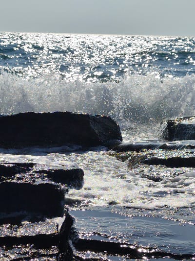 The image depicts a wave crashing against dark, rugged rocks at the edge of a body of water. The sunlight glints off the water's surface, creating a shimmering, sparkling effect, and also catches the spray from the wave. The rocks appear wet and dark, with the water flowing around and over them. The background shows the horizon where the water meets a hazy sky, suggesting an outdoor, coastal or lakeside environment. The scene appears to be located in the city of Saiyn, Kazakhstan.