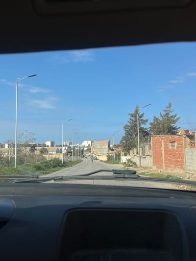 The image, captured from inside a vehicle through its windshield, depicts a street scene in Ain El Turk, Algeria, under a clear blue sky with scattered white clouds. The vehicle's dashboard and windshield wipers are visible in the foreground. A paved, two-lane road extends into the midground, curving slightly to the right, with a faint white center line. One individual is visible walking in the middle of the road, away from the camera, wearing light-colored attire.
To the left of the road, a vegetated verge precedes a series of buildings, some appearing completed multi-story structures, others seemingly under construction with visible concrete frames. Multiple modern streetlights with horizontal LED fixtures line this side. To the right of the road, the immediate area features sparse vegetation and a red brick building with exposed concrete elements, appearing unfinished or partially damaged. Further back, another incomplete concrete structure and a wall border the road. Several tall, slender coniferous trees are interspersed among these structures and additional streetlights. The environment suggests a mix of developed and developing urban or suburban areas.