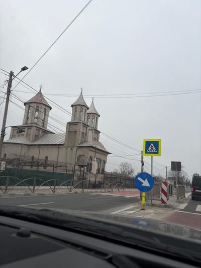 The image depicts a static street scene in Tuzla, Romania, viewed from inside a vehicle. The sky is uniformly overcast, indicating a lack of direct sunlight.

In the foreground, the lower portion of the image shows the dark dashboard and the bottom edge of a windshield. The scene beyond is primarily composed of a road, a prominent church, and various urban elements.

A multi-lane asphalt road runs horizontally across the midground. On the far right, a black van, possibly a Mercedes-Benz Vito, is visible, with a Romanian license plate starting "CT 19 M...". Adjacent to the road, on the right side, multiple traffic signs are present: a square yellow sign with a black pedestrian symbol indicating a crosswalk, and a round blue sign with a white arrow pointing diagonally downwards to the right, mounted on a yellow-and-black striped support. A red and white striped barrier is situated behind these signs. A section of the road in the immediate right foreground is marked with red paint.

On the left side of the road, a sidewalk is bordered by a decorative metal fence featuring repeating arched sections. Behind this fence stands a light-colored (beige/grey) Orthodox church, the primary subject in the background. The church features two prominent bell towers or domes with arched windows, each capped with a dark, possibly metallic, roof and topped with a cross. A third, smaller dome is partially visible behind the main structure. Wrought-iron gates provide access to the church grounds. Bare trees or vines on trellises are visible within the church compound, suggesting a dormant season. A utility pole with multiple overhead wires and a streetlamp is positioned on the left, partially obscuring the church. No individuals are visible in the scene.
