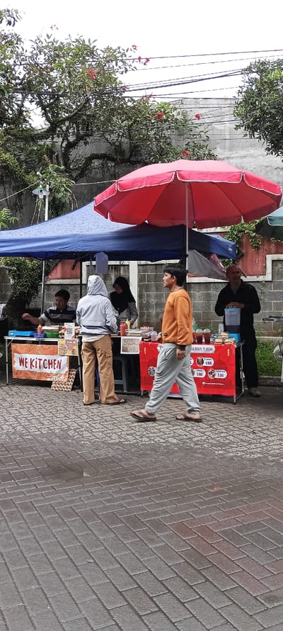 The scene is in front of a food stall in the city of Banjaran, Indonesia (latitude: -7.047, longitude: 107.58). The stall is under a blue canopy and a red umbrella. There are four people in the scene. Two are standing behind the food stall, and two are in front of it. One person is standing by a cooler, which contains a large container of ice. They are all wearing casual clothing. The stall features signs displaying the name “We Kitchen” and prices for food items. The stall is on a brick walkway. The background shows trees, a wall, and power lines. The sky is overcast.