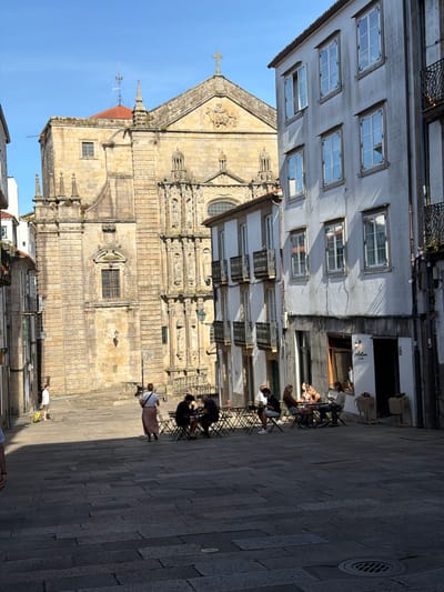 The image depicts a sun-drenched street scene in Santiago de Compostela, Spain. In the background, the imposing stone facade of a historical church, likely the Santiago Cathedral, dominates the view, with its intricate architectural details and a cross atop its roof. In the foreground, a narrow cobblestone street slopes downwards, lined with traditional European buildings. On the right, a multi-story building features a row of windows and balconies with wrought-iron railings. A cafe on the ground floor has outdoor seating with several patrons enjoying drinks and conversation. On the left, another building displays similar architectural elements. Several people are present: a person in a white shirt and shorts walks with a yellow suitcase, another person in a long skirt stands near tables, and a group of people are seated at the outdoor cafe. The sunlight casts distinct shadows across the cobblestone pavement, indicating a bright, clear day. The overall atmosphere is that of a vibrant European city square.