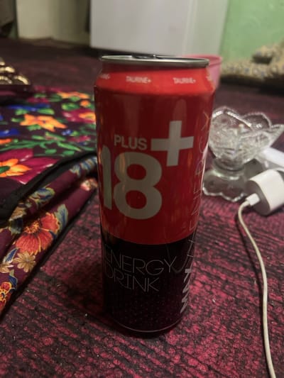 The scene depicts an indoor environment in Qoraobdol QFY, Uzbekistan. A cylindrical aluminum can, approximately 330ml-500ml in size, is centrally positioned on a dark red, woven fabric surface, likely a rug or carpet. The can's upper half is red, displaying "PLUS 18+" in large white lettering, with smaller "TAURINE+" text repeated above it. The lower half of the can is dark-colored, featuring "ENERGY DRINK" printed vertically in white text. The can's pull tab is open, indicating it has been unsealed. To the left of the can, a folded or layered piece of fabric with a distinct floral pattern, comprising red, orange, green, and blue flowers on a dark background, is visible. To the right of the can, a clear, cut-glass dish with an ornate, scalloped edge is present. Further right, a white power adapter is connected to a white charging cable, which extends towards the foreground. The background is out of focus, revealing indistinct lighter and darker surfaces. No individuals are present.