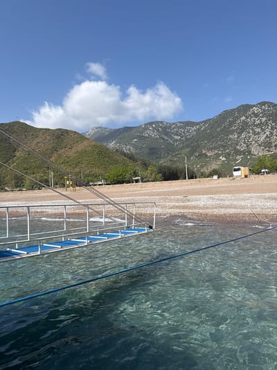 A landscape photograph taken at an angle shows a coastline in Kumluca, Turkey, with a rocky beach transitioning to clear water on the right. In the foreground, the blue and white railings of a boat or pier extend into the water. Along the beach, there are scattered vehicles and structures, possibly indicating a camping or recreational area.
Behind the beach, a mountain range rises, its slopes covered in greenery that transitions to exposed rock further up. A blue sky with scattered clouds is visible above the mountains. Power lines are also visible, stretching from the mountain area towards the beach.
