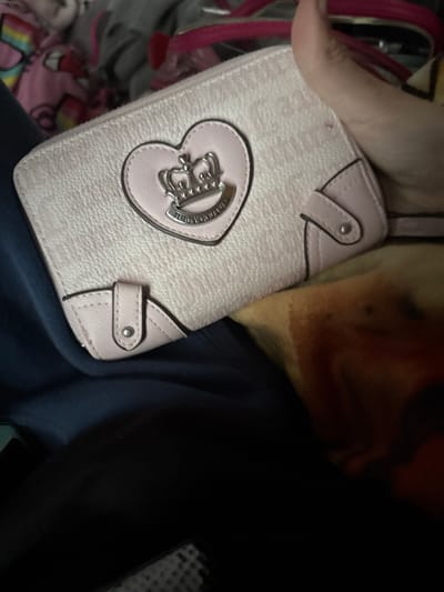 A pale pink leather wallet with a heart-shaped emblem featuring a crown and the text "JUICY COUTURE" is held by a person's hand. The wallet has a textured surface and pink trim with silver snap closures. The background is blurred and shows a pile of soft, colorful fabric, possibly clothing. The scene is indoors, with no clear indication of location within Gaylord, United States of America.