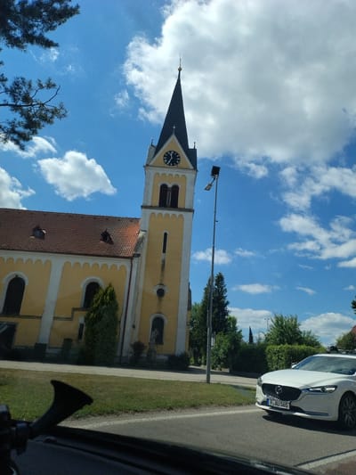 This is a slightly angled, eye-level, outdoor photograph taken on a sunny day with scattered white clouds in a bright blue sky. The scene features a prominent yellow church with a reddish-brown tiled roof and a tall, dark steeple topped with a gold finial. The church has arched windows and decorative white trim. A clock face with Roman numerals is visible on the steeple. A tall street light stands to the right of the church. In the foreground, a white car with a license plate reading "P AU 565" is driving on a paved road. The car has a roof rack. To the left of the church, a dark silhouette of a car's interior is visible, including a black horn-like object and a windshield. Green grass and trees are present in the background and along the side of the road. The photograph appears to be taken in the town of Bližná, Czechia.