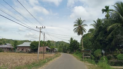 The image depicts a rural scene on a paved road in Blangrheuem, Indonesia, under a partly cloudy sky. On the left side of the road, a dry rice field is visible, with two houses constructed from brick and corrugated metal roofs. Utility poles with visible power lines are positioned along the road. The right side of the road features dense green vegetation, including several palm trees and other tropical foliage. A wooden structure, possibly a roadside stand, is also present. The road curves gently into the distance, leading towards a hilly landscape in the background. The overall atmosphere is one of a quiet, sunlit day in a tropical rural setting.