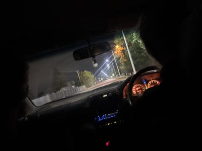 The image depicts the interior of a car at night, with the camera positioned to show the dashboard and the road ahead. The dashboard features illuminated gauges, a digital clock displaying "13:43", and a car radio with blue lights. Outside the car, streetlights line the road, and trees with orange foliage are visible. The scene suggests movement, as indicated by the blurred motion of the surroundings. The location is identified as Ptichnik, Russia.