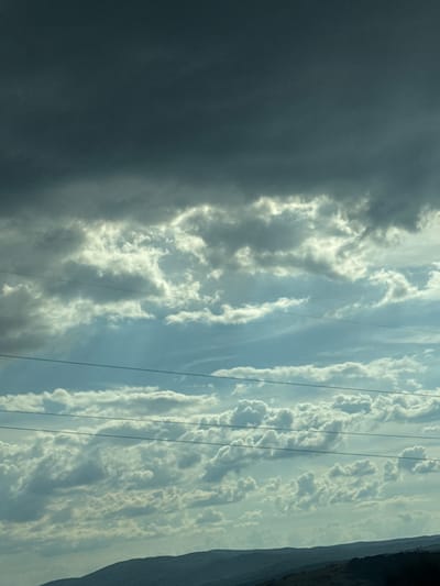 The image depicts a dramatic sky over a silhouetted landscape, likely Verinsko, Bulgaria. The sky is dominated by a variety of cloud formations. The upper portion of the image features dark, heavy storm clouds, appearing almost black and foreboding. Below these, lighter, wispy clouds are illuminated by sunlight, creating a contrast between darkness and light. Several thin, dark lines, presumably power lines, stretch horizontally across the middle of the frame. The lower part of the image shows the gentle, rolling contours of a dark, mountainous or hilly landscape. The overall impression is one of an approaching or passing storm, with a sense of vastness and natural power. There are no people or animals visible in the image.