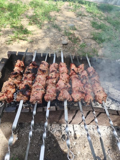 The image shows skewers of meat cooking over coals in a brazier, likely a shashlik. The brazier is situated outdoors on dirt ground with sparse vegetation, suggesting a rural or semi-rural setting. The meat appears to be partially cooked, with some charring on the edges. Smoke rises from the coals and the meat. The background is out of focus, showing more vegetation and a wall or fence. The scene is indicative of an outdoor barbecue or cooking event.