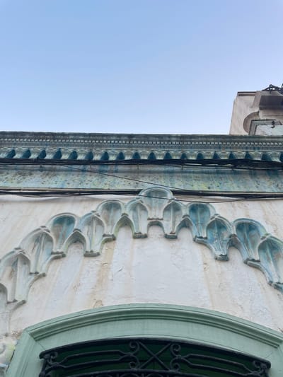 This is a low-angle, exterior shot of a building's facade in La Línea de la Concepción, Spain. The sky is clear and a pale blue, taking up the top half of the image. The building features architectural details with a distressed, aged appearance.

The upper part of the facade has a band of decorative molding with a textured, almost serrated edge, painted in a faded teal color. Below this, a horizontal line of dark electrical wires runs across the wall. Further down, a series of arch-like decorative elements are prominently featured. These elements are painted in a light cream color, with accents of pale teal and hints of yellowing and water stains, suggesting age and weathering. The texture of the wall is rough and uneven.

At the bottom of the image, the curved top of a window or doorway is visible, framed by a green-painted border. Below this frame, a dark, ornate wrought-iron grille is present, partially obscuring what lies beyond. The overall impression is of an old building with intricate, historical architectural features that show signs of wear and tear.