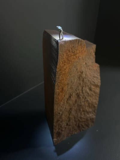 The image presents a studio-lit miniature diorama, conceptually set in the city of Galatina, Italy. The central subject is a single, small human figurine, approximately 2-3 cm tall, wearing light-colored attire. The figure is positioned on the uppermost edge of a large, vertically oriented block of rough, reddish-brown material, resembling coarse-grained wood or sandstone. The figure is bent forward, with its head and torso angled downwards, appearing to observe or investigate the immediate area below its feet on the block's surface. A localized, intense white light source, emanating from the figure's upper body, illuminates this specific point. The block exhibits a textured, uneven front face and a smoother, darker, striated left side. It rests on a flat, dark surface, casting a defined shadow. The background is uniformly dark gray, with a faint diagonal line visible in the lower-left, suggesting a controlled photographic environment.