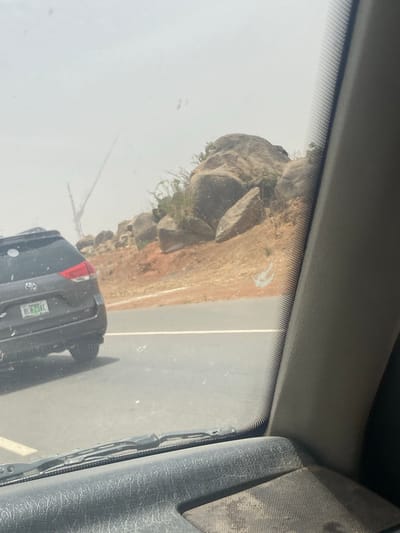 The image shows a view through a car windshield, likely from the passenger seat. Outside, there is a rock formation adjacent to a paved road. A black car is partially visible in the lower part of the frame, appearing to be moving on the road. The location can be inferred to be Zangon Aya, Nigeria, based on the license plate on the car. The environment appears dusty or hazy.