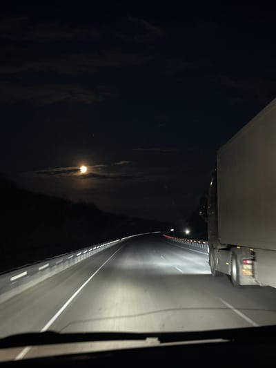 The image shows a night view from a high position, possibly a bridge, overlooking a road. The right side of the image is dominated by a concrete barrier and a multi-lane road illuminated by bright overhead lights. White lines mark the lanes. The left side is dark with indistinct cloud formations and a distant, dim light source, possibly the moon. The viewpoint is angled downwards. No people or subjects are directly visible. The location of Arkhara, Russia, is not confirmable from the visual content alone.