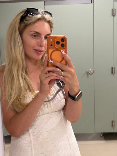 A blonde woman is taking a selfie in a mirror. She is wearing a white dress and black sunglasses. The background appears to be a locker room, with mint green lockers visible. She is holding an orange phone, and her nails are white with black hearts. She is also wearing a gold watch and a ring. The lighting suggests it could be daytime, and the setting seems to be indoors. The woman's expression is neutral, and she is looking directly at the camera. The word "vogue" is visible on the sunglasses.