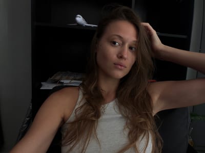 A young woman with light brown hair and fair skin is positioned upside down in the image. She is wearing a light-colored tank top and a delicate necklace. Her arms are raised, with one hand resting near her head. Below her, a dark shelving unit is visible. On one of the shelves, there is a model airplane. The background is a mix of dark and light tones, suggesting an indoor setting. This image does not contain information to ascertain that the location is in Kerobokan Kelod, Indonesia.