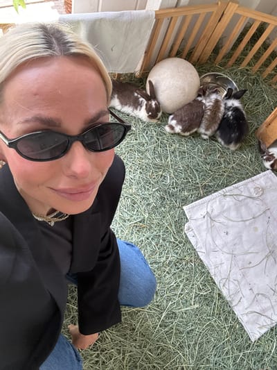 The image appears to be taken from a first-person perspective, looking down at a rabbit pen in Solana Beach, California. Several small, mixed-color (brown/white and black/white) rabbits are visible near a metal bowl and a rock. A white cloth is laid out in the pen, which is filled with hay. A person wearing black sunglasses, a black blazer, blue jeans and a gold necklace is in the foreground. The pen appears to be partially constructed of wood.