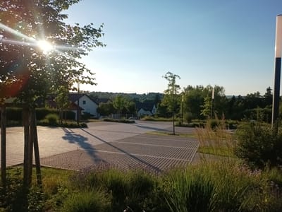 A sunny, wide-angle, eye-level outdoor shot captures a paved parking area with a grass and foliage border. The sun's rays pierce through the leaves of a tree on the left side of the frame, creating a lens flare. In the background, houses and trees are visible under a clear blue sky. The parking area is composed of a grid-like pavement, with some sections featuring a honeycomb pattern. The foreground is filled with lush green grass and various plants, including tall, feathery grasses and small purple flowers. A tall, modern street lamp stands on the right side of the image, and another is partially visible on the left. The scene suggests a residential or communal area in Oberviechtach, Germany, likely during a warm, clear day. No people are present in the image.
