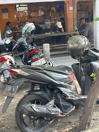 In Kludan, Indonesia, a street-side food stall is captured with several individuals engaged in activities. The foreground is dominated by parked motorcycles. One motorcycle, dark gray, has a license plate reading "N 2480 HJ" and "12-26". Another motorcycle, black, displays a license plate "W 3056 NEH" and "05-20". A third motorcycle, silver, has a partial license plate visible as "4016 VN" and "09-26". A green, white, and red helmet rests on the handlebars of a motorcycle.
Inside the stall, a person wearing a white cap is seated, leaning forward with their head down. Another person, wearing a dark jacket, is seated opposite them, smoking. To their right, another individual is seated. Behind the counter, another person is visible, facing away from the camera. The stall itself is stocked with various goods, including bottles of drinks and what appear to be packaged items. The environment suggests a casual gathering, possibly during a break or while waiting.