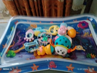 A baby's water play mat is shown, filled with water and featuring a colorful underwater scene with various toys. The toys include a pink pig with a blue hat, a yellow moon-shaped rattle, a turquoise ball with holes, a yellow toy with a face, and other small, brightly colored plastic items. The mat itself is transparent with a blue border and printed with images of starfish, crabs, and octopuses. The background shows a blurred curtain. The timestamp "12.07.2026 20:10" and "REDMI 15" are visible in the bottom corners. There are no people visible in the image. The scene appears to be indoors, likely in a home environment. The city of Айет, Kazakhstan, is not depicted or suggested in the image.
