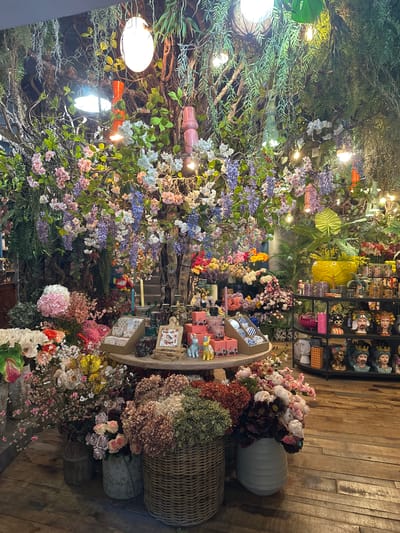 This is a photograph taken inside a flower shop in Oviedo, Spain, likely during daylight hours, though the artificial lighting within the store is prominent. The scene is densely packed with a variety of artificial and preserved flowers and plants, creating a lush and slightly whimsical atmosphere.

In the foreground, a wooden floor with visible grain and planks leads into the space. To the left, large baskets and vases overflow with dried and artificial flowers in muted tones of green, brown, and pink. One large wicker basket in the center-right foreground contains a dense arrangement of dried hydrangeas in shades of pink and brown. To its left, a vase holds a cluster of pastel pink and white roses. Another tall vase on the left contains a mix of pink and white artificial flowers.

Dominating the mid-ground is a large, tree-like structure with branches that extend upwards and are adorned with an abundance of artificial flowers in shades of pink, white, purple, and blue. Hanging vines and greenery cascade from the upper branches. Several round, white pendant lights and orange cylindrical lights are suspended from the ceiling, casting a warm glow.

A round display table is situated in front of the floral tree, showcasing a collection of small boxes, ceramic figurines (including a white rabbit), and decorative items. Behind this table, a shelf unit displays an assortment of decorative ceramic busts and jars with colorful patterns.

The background of the shop is filled with more plants, flowers, and shelving units, suggesting a substantial inventory. The overall impression is one of an abundant and elaborately decorated retail space specializing in floral and decorative items. There are no people visible in the photograph.