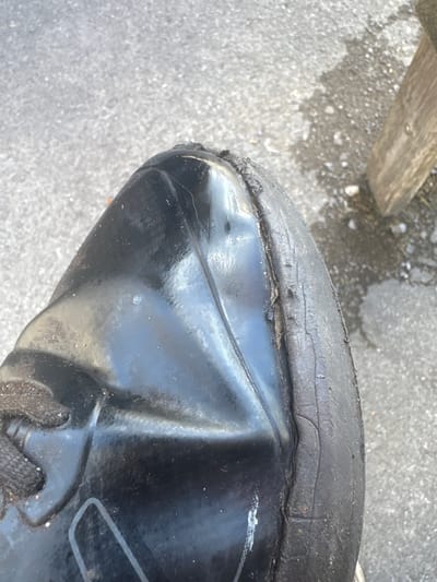 A close-up, high-angle shot shows the worn tread of a black shoe, likely a sneaker or athletic shoe, resting on a paved surface. The shoe's rubber sole is significantly deteriorated, with visible cracks and fraying along the edge, exposing the inner layers. The upper portion of the shoe is made of a glossy black material, possibly synthetic leather or plastic, showing scuffs and minor damage. The shoe is positioned on a grey asphalt or concrete ground, which appears damp in places, suggesting recent rain or a wet environment. In the background, a wooden post is partially visible on the right side, and the texture of the ground indicates an outdoor urban setting. The scene is devoid of people, focusing solely on the damaged footwear. The location is specified as Tewkesbury, United Kingdom.