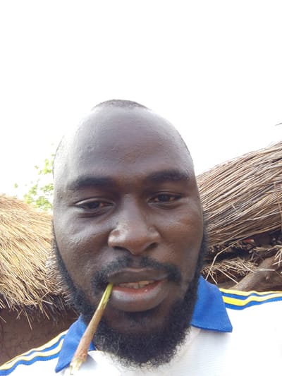A close-up selfie of a Black man with a bald head, a beard, and a short stem in his mouth. He is wearing a white shirt with blue and yellow trim. Behind him is a thatched structure, possibly a roof or wall, with visible branches and debris. The setting appears to be outdoors. There is no discernible information about Mulke Barikin Sayejo, Nigeria to determine the location.