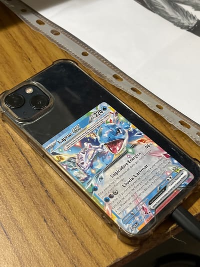 The image shows a smartphone in a transparent case with a Pokémon card placed inside, behind the phone. The card features the Pokémon Lapras ex, with Spanish text on it. The phone is resting on a wooden surface, with a white sheet of paper with holes punched on its left side. A black cable is connected to the phone. There is no information to determine that the location is in the city of Bahía Blanca, Argentina.