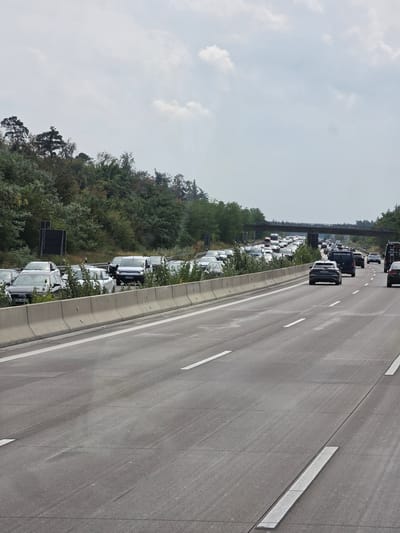 This image captures a traffic jam on a highway, likely in St. Leon, Germany, given the context. The scene is dominated by a multi-lane highway with a significant number of cars, trucks, and vans. The vehicles are mostly stationary or moving very slowly, indicating a traffic congestion. The highway is bordered by a concrete barrier on the left and a dense forest on the right. Overpasses are visible in the distance, suggesting a complex road network. The sky is overcast with some clouds. The overall impression is one of a typical highway scene during peak hours or due to an incident.