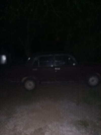 The image shows a dark, blurry view of a dark-colored car parked outdoors at night. The car appears to be a sedan with visible wheels and side mirrors. The background is very dark, with some faint hints of foliage or trees above the car. The ground in front of the car is visible and appears to be rough, possibly gravel or dirt. A faint light source is visible on the left side of the image, which may be a building or another light. The image is low quality and out of focus, making precise identification of details difficult. The location is identified as Yampil, Ukraine.