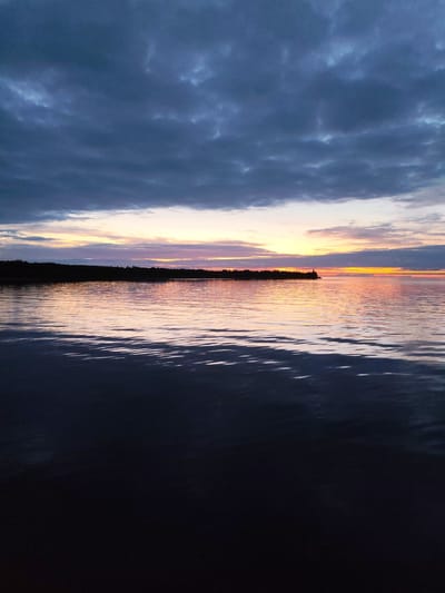 A serene sunset over a body of water near Shcheleiki, Russia, paints the sky with hues of orange, pink, and purple, contrasting with the dark, imposing clouds above. The water reflects the colorful sky, creating shimmering ripples on its surface. A distant, dark silhouette of a treeline stretches across the horizon, punctuated by a small, lit structure, likely a lighthouse or a beacon, on a point of land jutting into the water. The foreground of the water is almost entirely dark, with only faint glints of light suggesting its depth. The overall atmosphere is peaceful and tranquil, with the natural beauty of the sunset dominating the scene.
