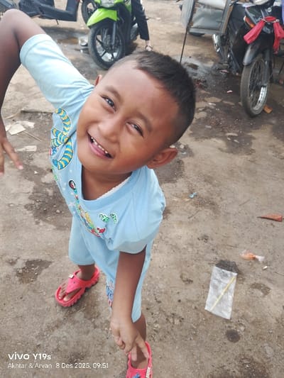 A young boy in a light blue short-sleeved outfit and red sandals stands on a dirty, cracked surface. He has short, dark hair and a wide, toothy smile. His arms are extended, one angled upwards and the other pointing down. In the background are motorbikes, and on the ground near the boy are a small bag with dark contents and a discarded wrapper. A smartphone watermark is at the bottom left.