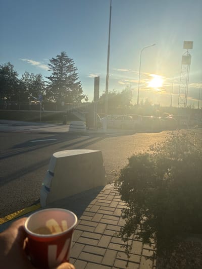 The photograph captures a sunny outdoor scene, likely at a rest stop or service area, given the presence of parked cars, a parking lot, and concrete barriers. The sun is low in the sky, casting a warm, golden light across the scene and creating long shadows on the asphalt and paved walkway. In the foreground, a hand holds a red and white paper cup filled with what appears to be a beverage and some solid contents. To the left of the cup, a concrete barrier with blue and white stripes is visible, and further back, another larger, curved concrete barrier is prominent. The background features a playground with a small wooden structure and a parking symbol, several trees, including a tall, dark evergreen, and a series of parked vehicles. A tall metal tower with a sign is visible in the distance, along with streetlights. The sky is bright blue with scattered clouds, and the overall atmosphere suggests late afternoon or early evening. The image appears to be taken in Kamenice, Czechia, based on the prompt's information.