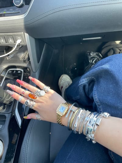 The image displays the interior of a vehicle, focused on the left hand and forearm of an individual, with portions of their lower body and the car's interior visible. The individual is wearing dark blue denim jeans. Their right foot, clad in a light-colored athletic shoe, is partially visible on the passenger-side floor. A crumpled black object, possibly fabric or a bag, rests near the shoe.
The vehicle's interior features a dark dashboard with visible stitching. On the left, parts of the center console and dashboard controls are discernible, including buttons labeled "SYNC" and "A/C," along with seat heater controls (L3, L5). A white USB cable is plugged into a port beneath these controls. Bright sunlight illuminates the scene from the upper right, creating shadows.
The individual's left hand has long, almond-shaped artificial nails, decorated with varied patterns and colors, including red glitter, yellow, and orange. The hand is heavily accessorized with rings on all fingers except the thumb. The middle finger prominently features a large oval, amber-colored stone set in a silver-toned ring. Other rings include multiple silver-toned bands and designs, one on the ring finger depicting a red "evil eye." The left wrist is adorned with a gold-toned rectangular watch, featuring a light-colored face surrounded by small clear stones on the bezel, and a gold-toned metal link band. Below the watch, a collection of bracelets is stacked on the forearm. These include a gold-toned "nail" design bracelet, a silver-toned "nail" design bracelet, several plain and textured silver-toned bangles, a thin silver-toned chain bracelet with small clear stones, and a bracelet with a heart-shaped charm.
The specific location within the city of Jisr az-Zarqa, Israel, is not discernible from the visual information provided in the image.
