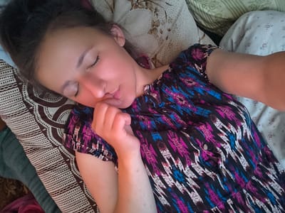 A young woman is captured in a selfie while lying down. She is wearing a colorful, patterned top with shades of black, blue, and pink. Her eyes are closed, and her head is resting on a patterned pillow. Her right hand is brought up to her face, with her fingers lightly touching her chin and mouth. The setting appears to be indoors, possibly a bedroom, with other pillows and bedding visible in the background, suggesting a comfortable and relaxed atmosphere. The lighting in the image is warm, with a slight reddish cast, and illuminates her skin and hair. The overall impression is one of peaceful rest or sleep. The location context provided is Anastasivka, Ukraine. There is no visible text in the image.