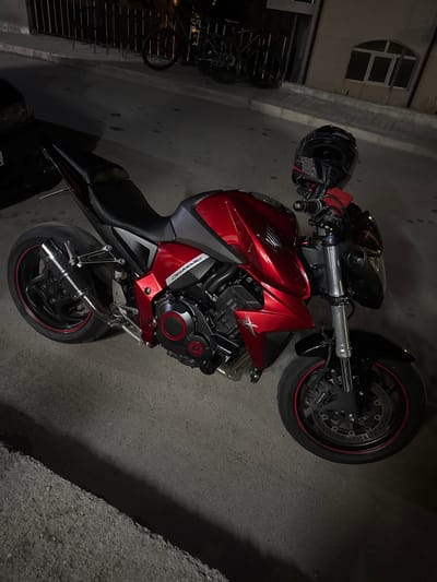 A red and black Honda motorcycle is prominently displayed, parked outdoors at night in Kraishte, Bulgaria. The image is taken from an elevated, slightly rotated angle, showcasing the left side of the vehicle. The motorcycle features a glossy red fuel tank and side fairings, contrasted with a black seat, frame, and wheels. Text such as 'HONDA' is visible on the fuel tank, and 'EXTREME' is present on a side fairing, along with a smaller 'CB' inscription near the seat.
A black motorcycle helmet, with some patterned details, rests on the handlebars. The front wheel is equipped with disk brakes, and 'TOKICO' is visible on the brake caliper. Both the front and rear wheels have striking red pinstriping along their rims. An aftermarket 'DELKEVIC' exhaust system, partially covered in a carbon fiber-like wrap, is prominent on the right side of the bike. The engine casing features black and red accents.
The scene is dimly lit by artificial light, creating distinct shadows on the dark asphalt ground where the motorcycle is parked. In the background, a light-colored building with a distinctive arched, segmented window is partially visible. Below the window, a fence or railing and what appears to be part of a bicycle are also discernible. The overall atmosphere is quiet and nocturnal, highlighting the details of the well-maintained motorcycle in an urban or semi-urban setting. No people are visible.